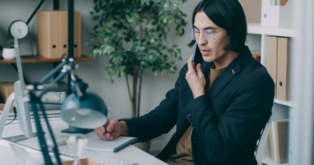 Man in glasses takes notes while talking on his phone at a desk.